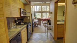 A compact kitchen with wooden cabinets, a microwave, a stove, and a dishwasher. There's a dining area with a table and chairs, and a view of the outdoors through large windows. - Slopeside 2771 (Keystone)