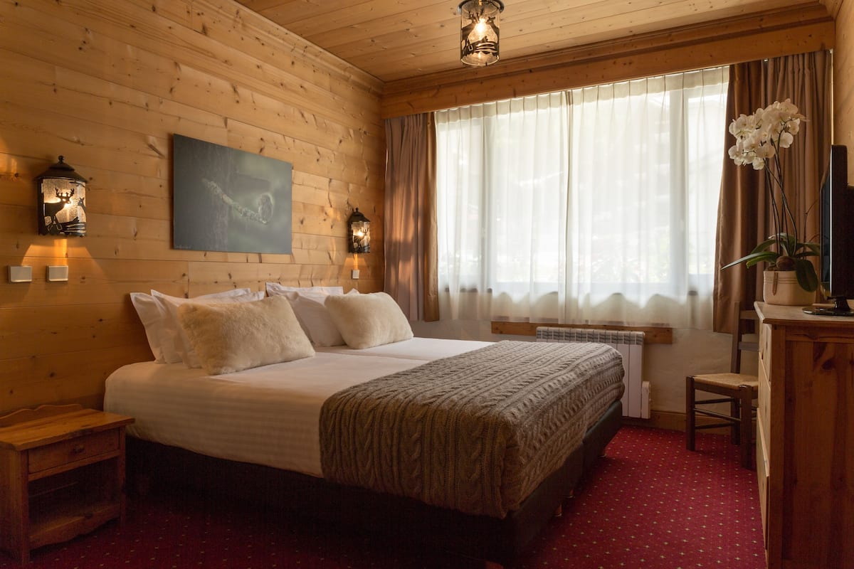 Ein schlafgemäch mit Holzvertäfelung, einem großen Bett, zwei Wandlampen, einem Fenster mit Vorhängen und einem kleinen Schreibtisch mit Stuhl.
