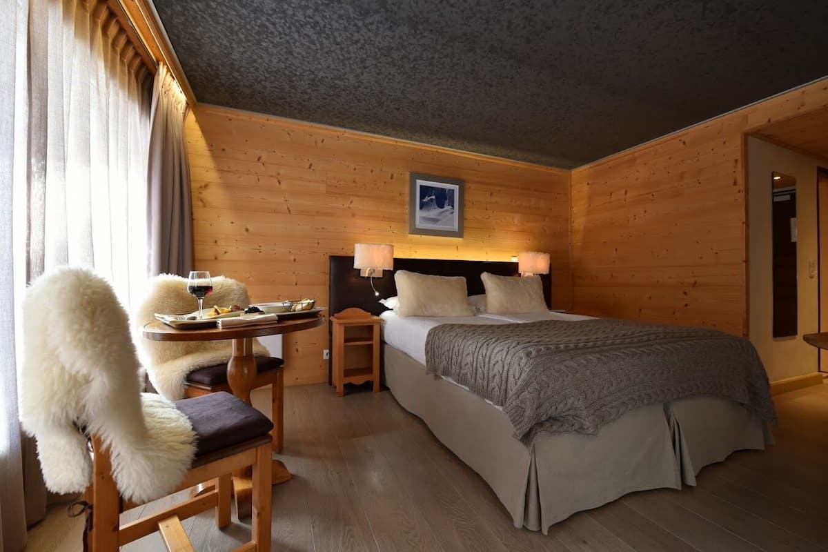 Ein gemütlicher Holzraum mit einem Bett, einem kleinen Tisch mit Stuhl und einer Lampe.