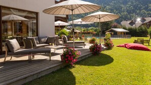 Een terras met buitenmeubilair en parasols, omgeven door een verzorgd gazon en een berg op de achtergrond.