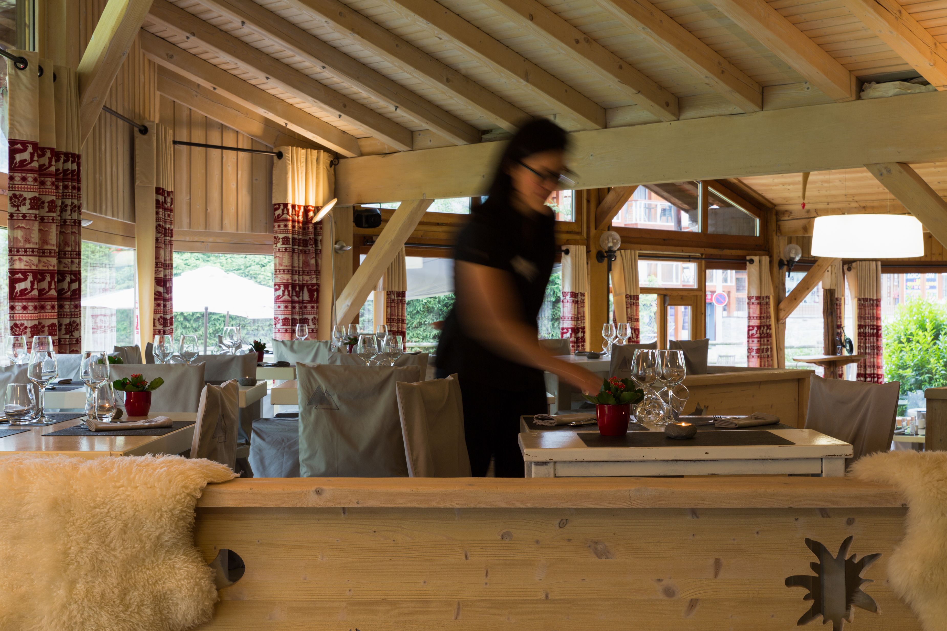 Un restaurant avec des poutres en bois, des tables dressées avec des verres à vin, et un serveur.