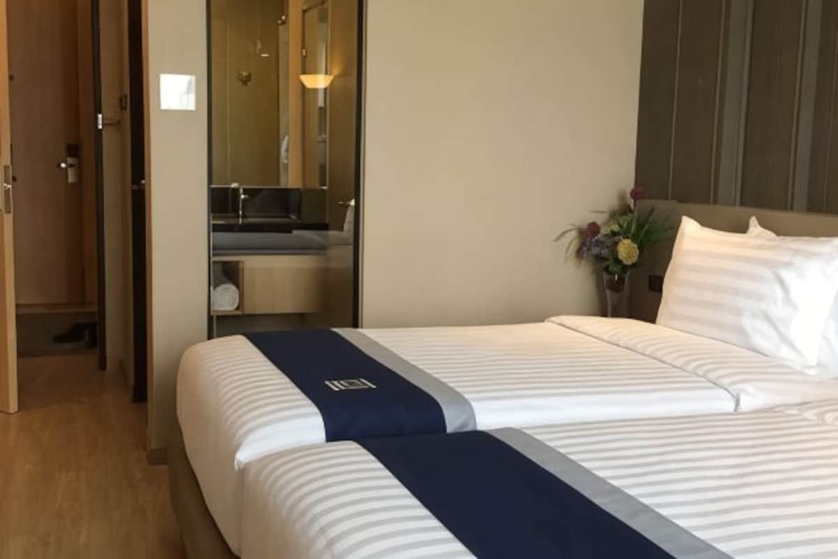 Una camera d'albergo con due letti, un bagno con uno specchio e una piccola pianta sul comodino.