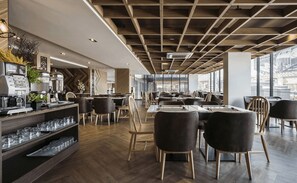 A modern restaurant with wooden flooring, black chairs, and a bar area with glasses and a coffee machine. - Zayn Hotel Bangkok (Bangkok)