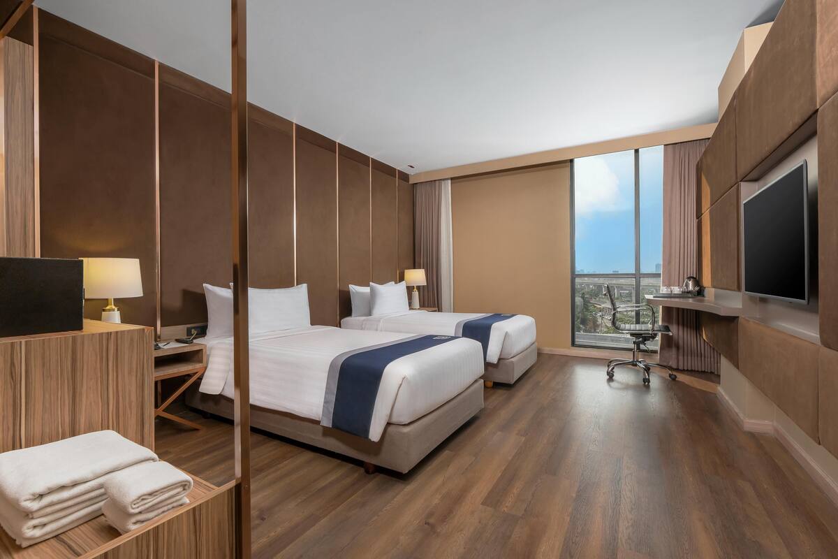 Een moderne hotelkamer met twee bedden, een bureau, een stoel, een televisie en een groot raam met uitzicht op de stad.