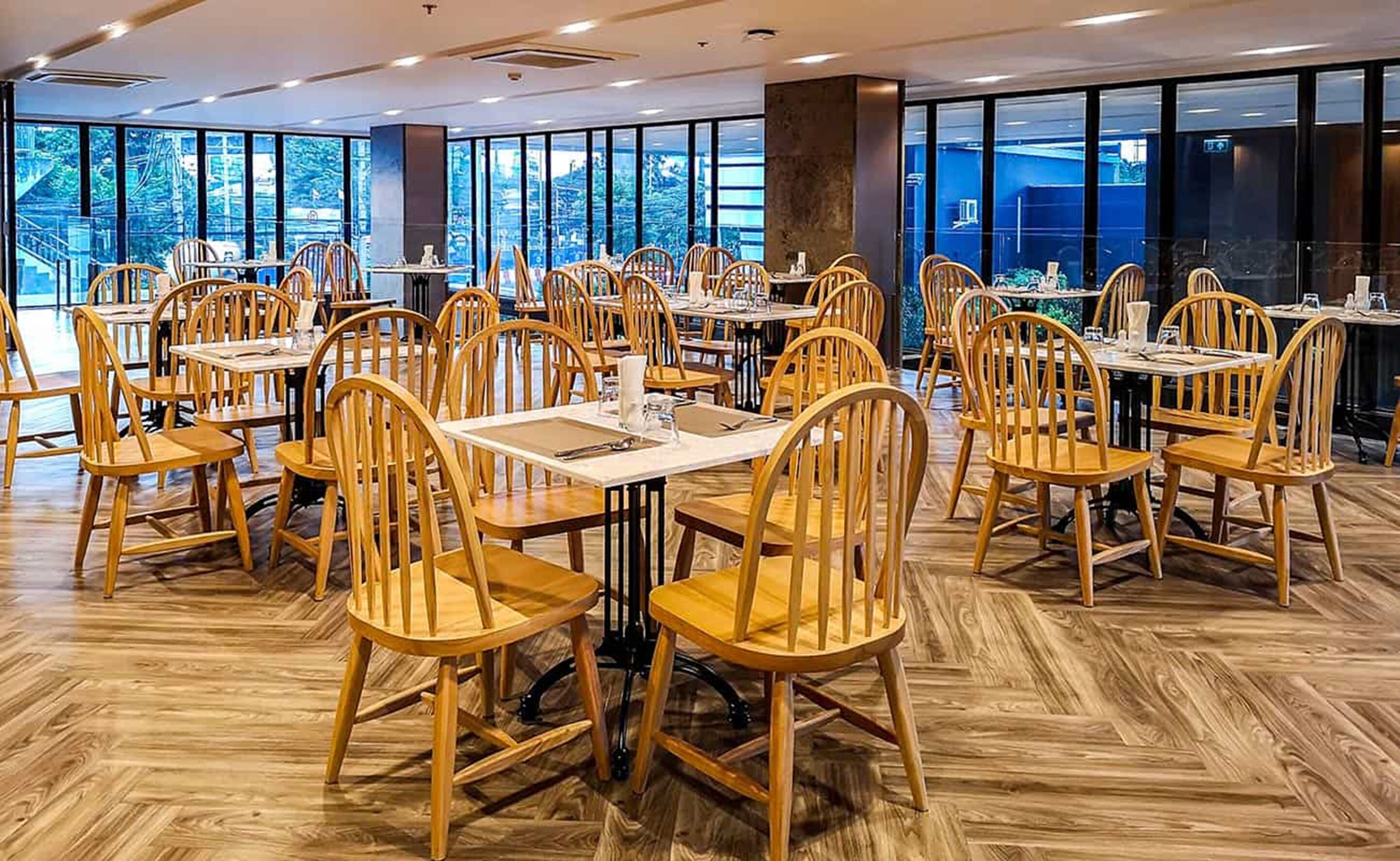 A spacious dining area with wooden chairs and tables, large windows offering a view of the outdoors, and a polished wooden floor.