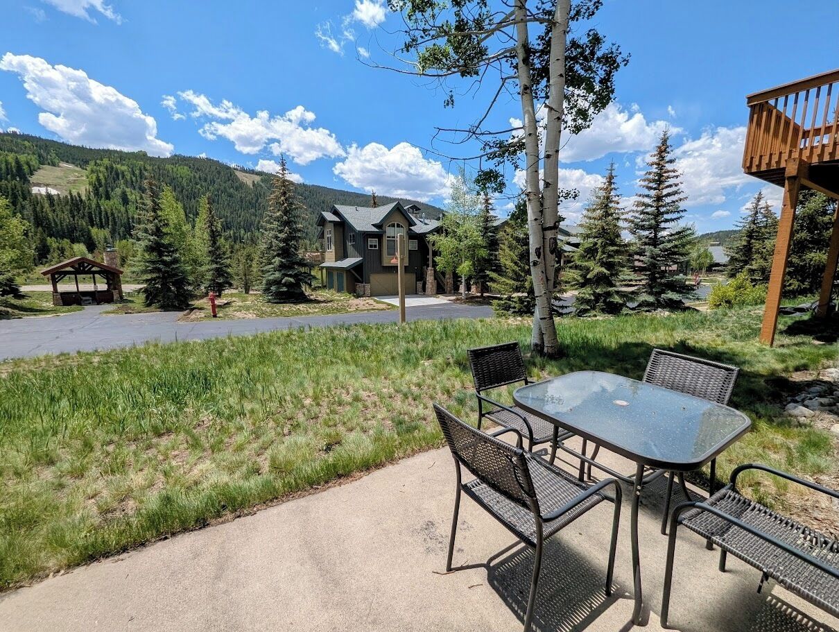 A patio with a glass table and chairs, surrounded by a grassy area and trees.