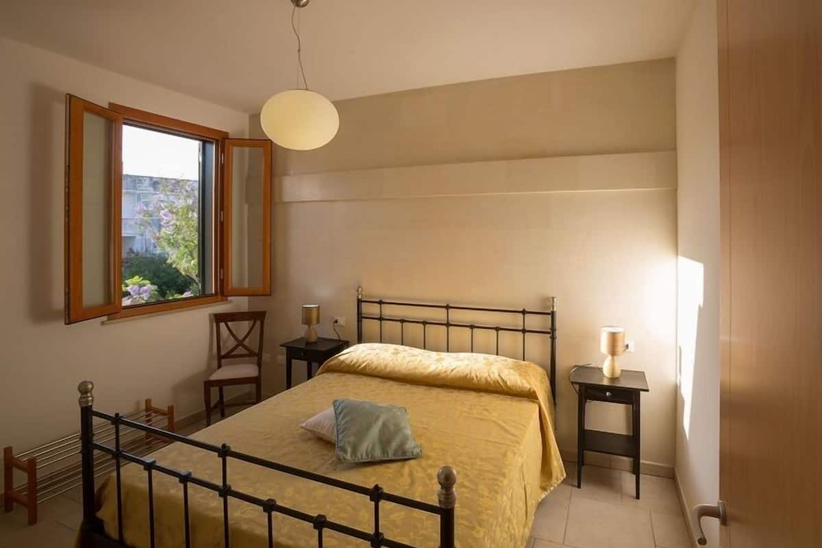 Une chambre à coucher avec un lit, une table de chevet, une chaise, une fenêtre et un miroir.
