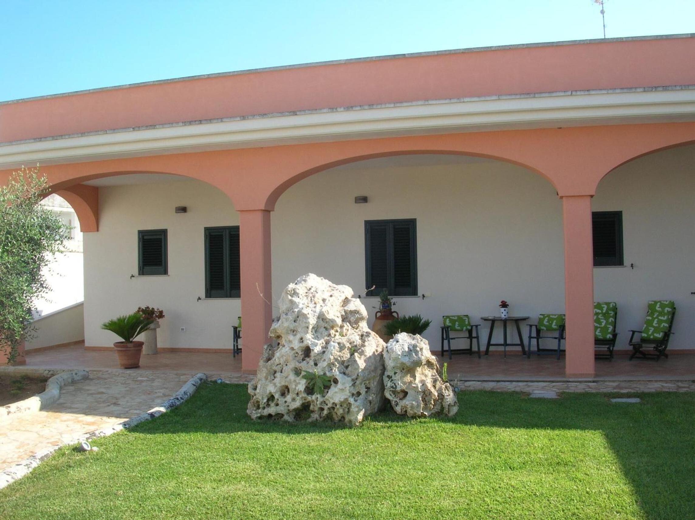 A building with a pink facade, three arched covered areas, outdoor seating, and a rock formation in the foreground.