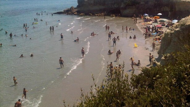 A crowded beach with people swimming and sunbathing, a rocky cliff, and umbrellas on the sand. - Casina dei Nonni (Carpignano Salentino)