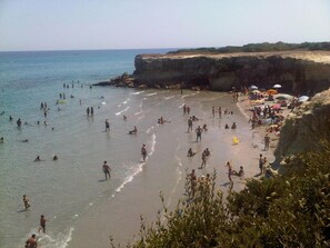 Una playa concurrida con gente nadando y tomando el sol, un acantilado rocoso y sombrillas en la arena.
