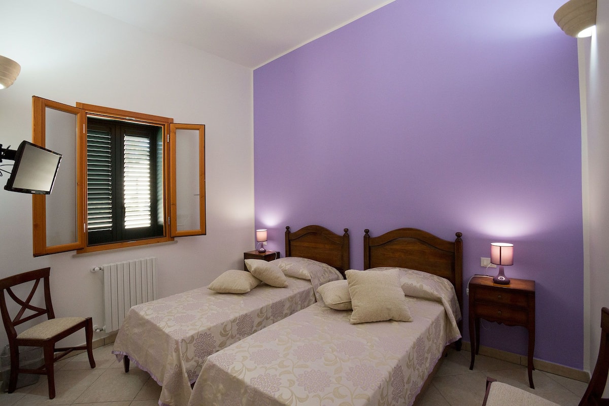 Une chambre avec deux lits, une chaise, un radiateur, une fenêtre avec des stores et des lampes de chevet.