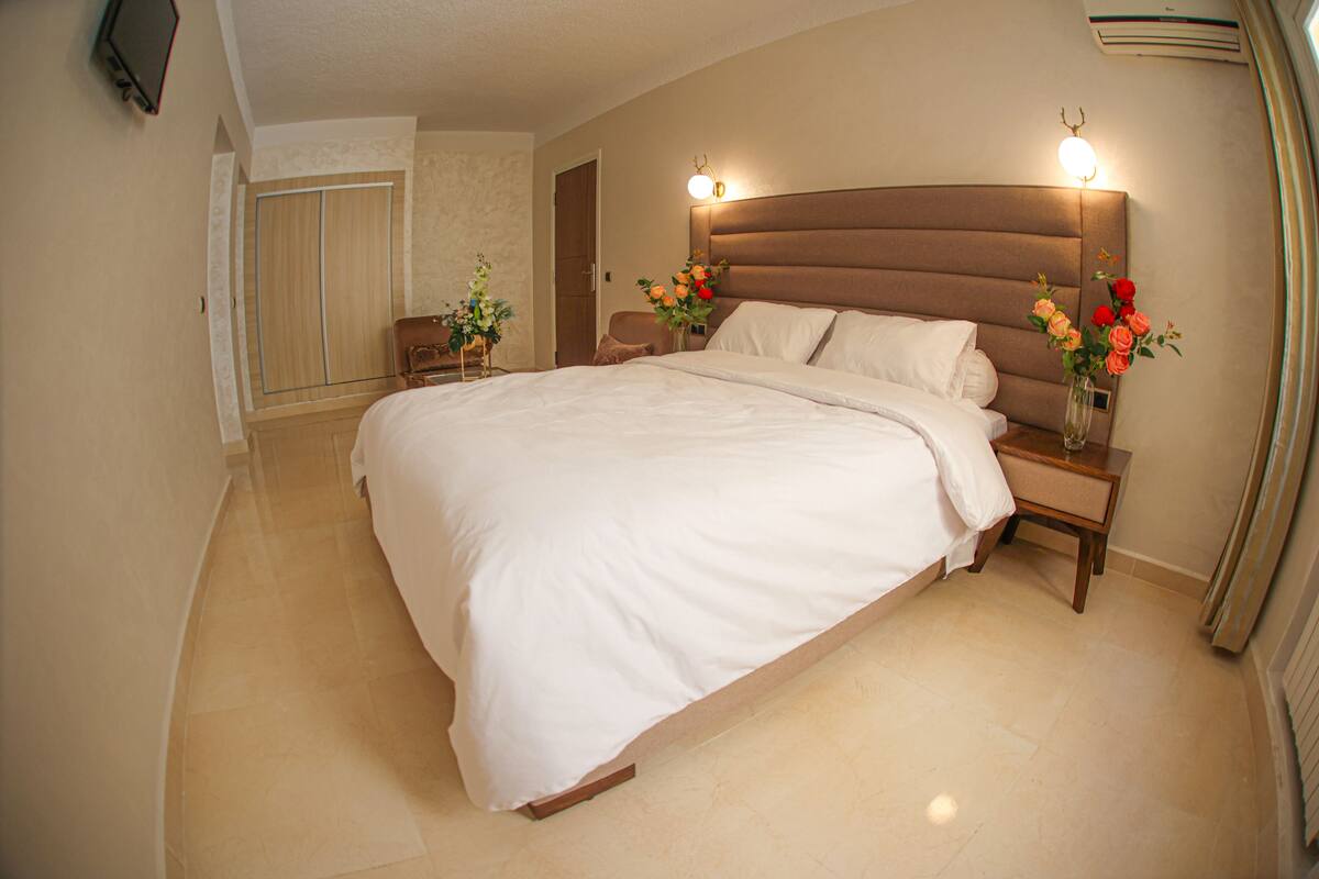 A bedroom with a large bed, a nightstand with a vase of flowers, a wooden wardrobe, and a television mounted on the wall.