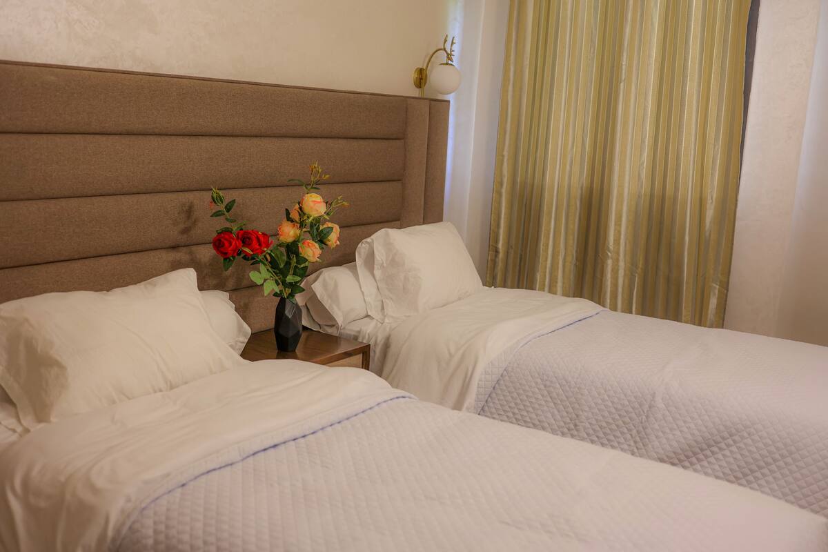A hotel room with two beds, a nightstand with a vase of flowers, and a window with curtains.