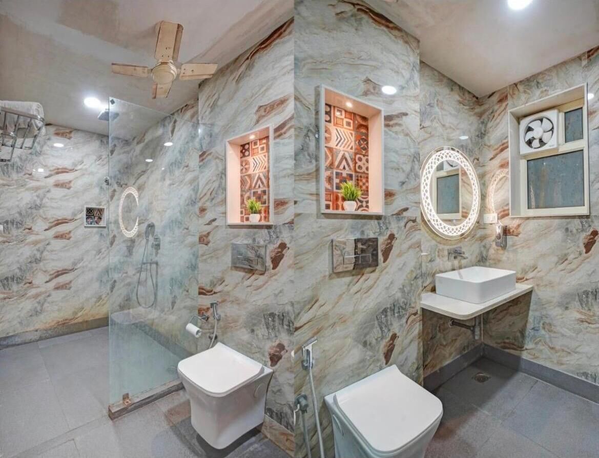 Un baño con paredes de mármol, cabina de ducha de vidrio, dos lavamanos, ventilador de techo y repisas decorativas en la pared.