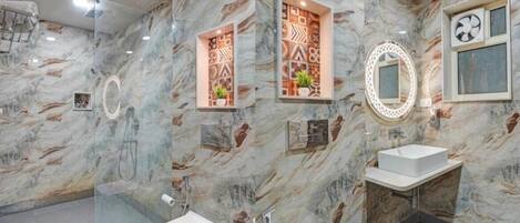 Un baño con paredes de mármol, cabina de ducha de vidrio, dos lavamanos, ventilador de techo y repisas decorativas en la pared.