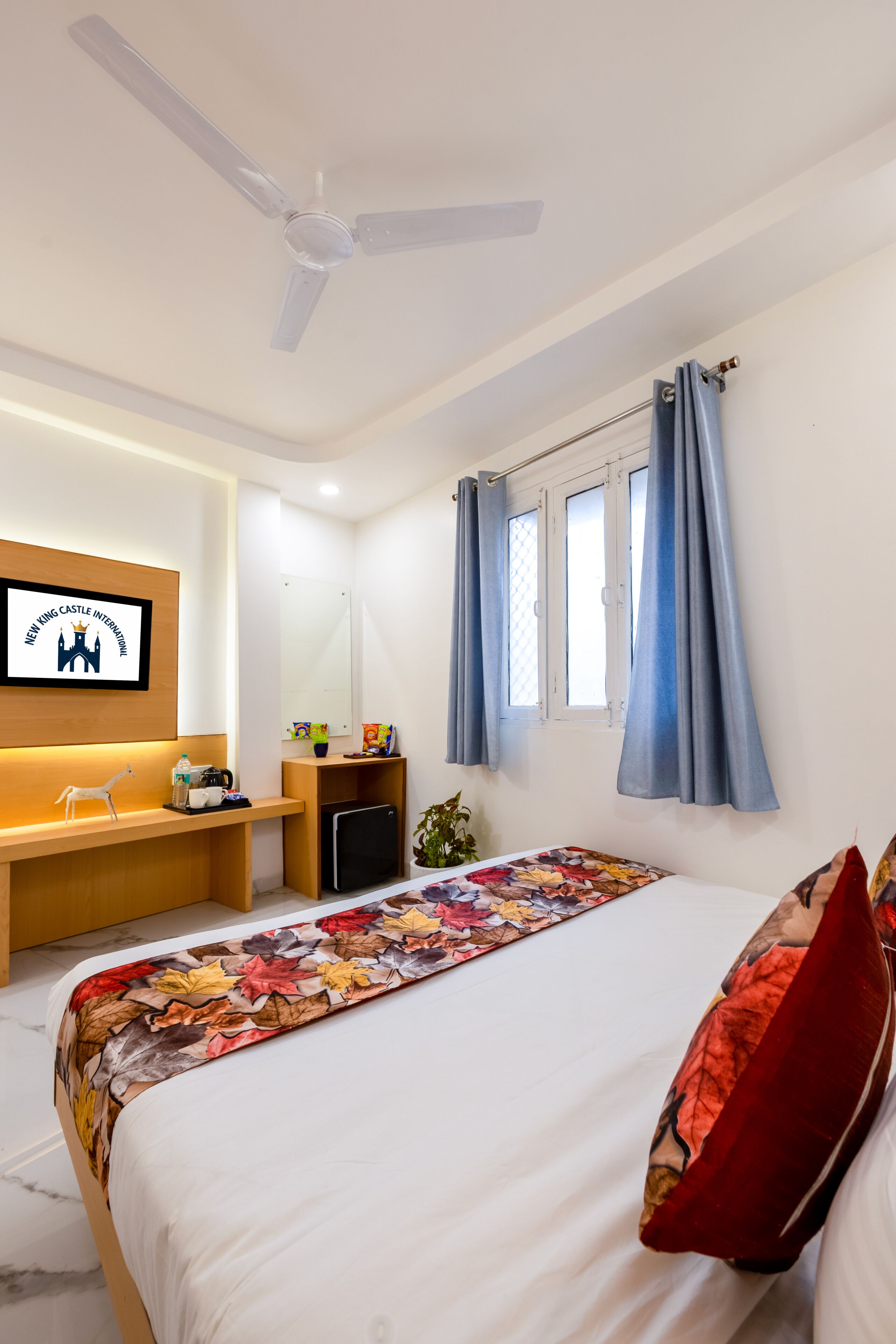 A hotel room with a bed, a desk, a chair, a ceiling fan, a window with curtains, and a framed picture on the wall.