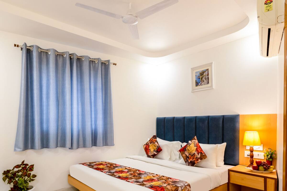 Ein Hotelzimmer mit einem Bett, einem Deckenventilator, einem Fenster mit Vorhängen, einer Nachttischlampe und einer wandmontierten Klimaanlage.