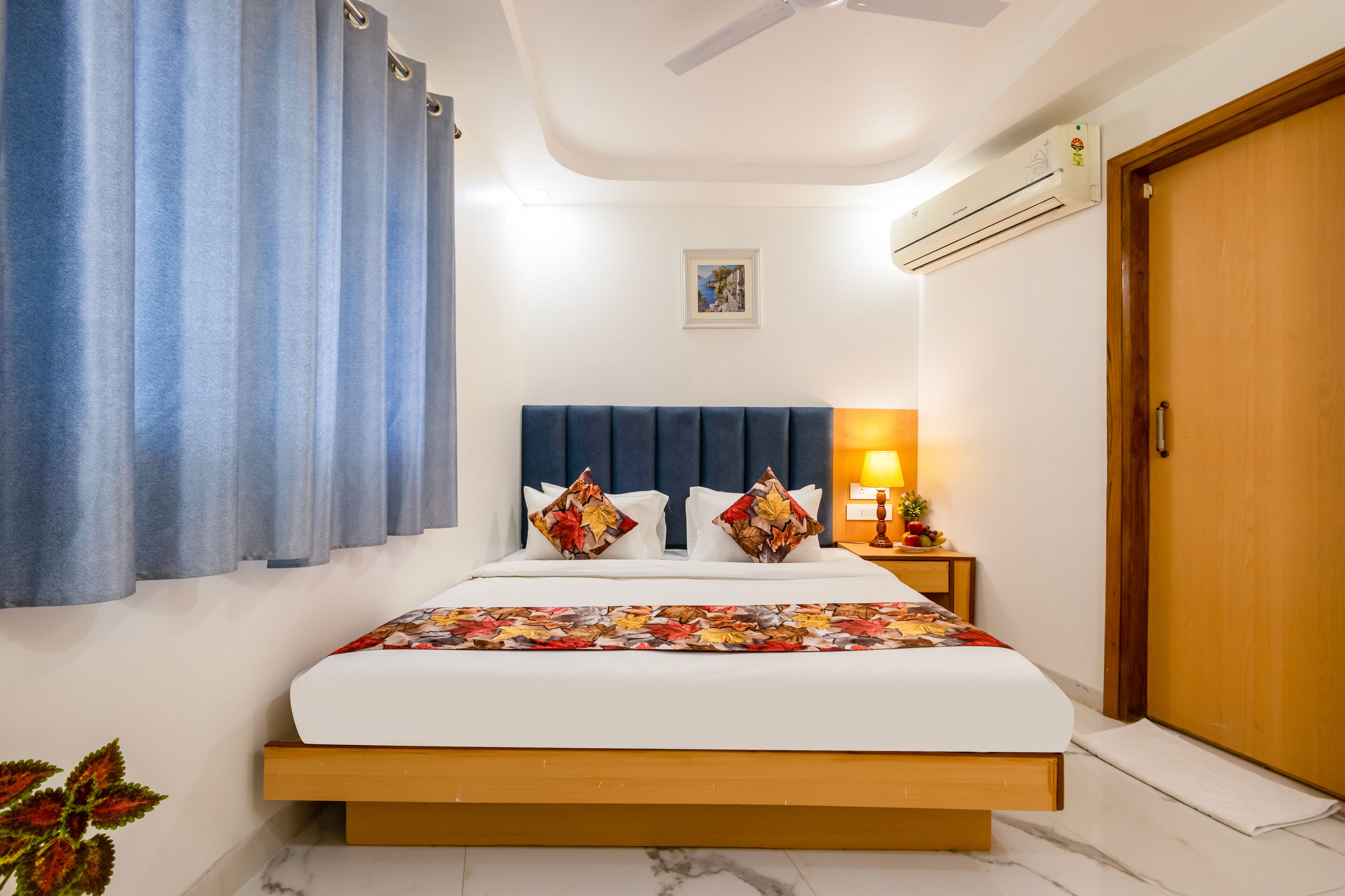 A bedroom with a bed, curtains, a nightstand, a lamp, an air conditioner, a door, and a picture on the wall.
