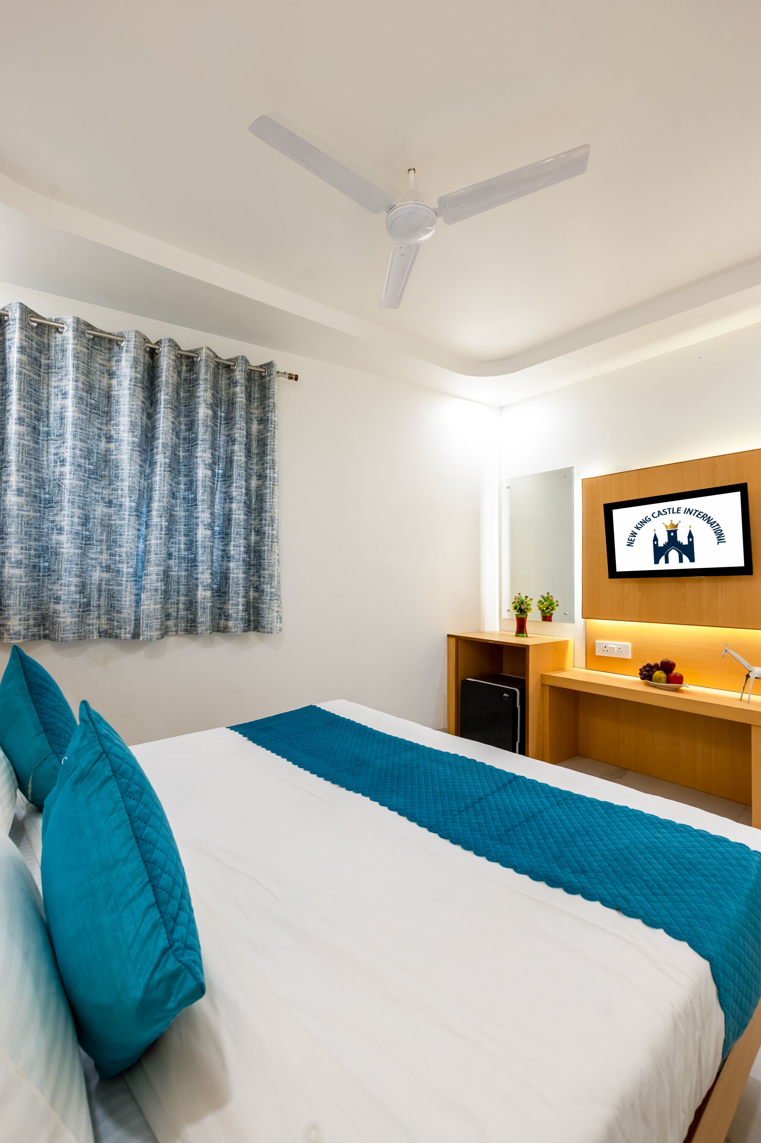 A hotel room with a bed, a ceiling fan, a desk with a computer, a mirror, and a wall-mounted TV displaying a castle logo.