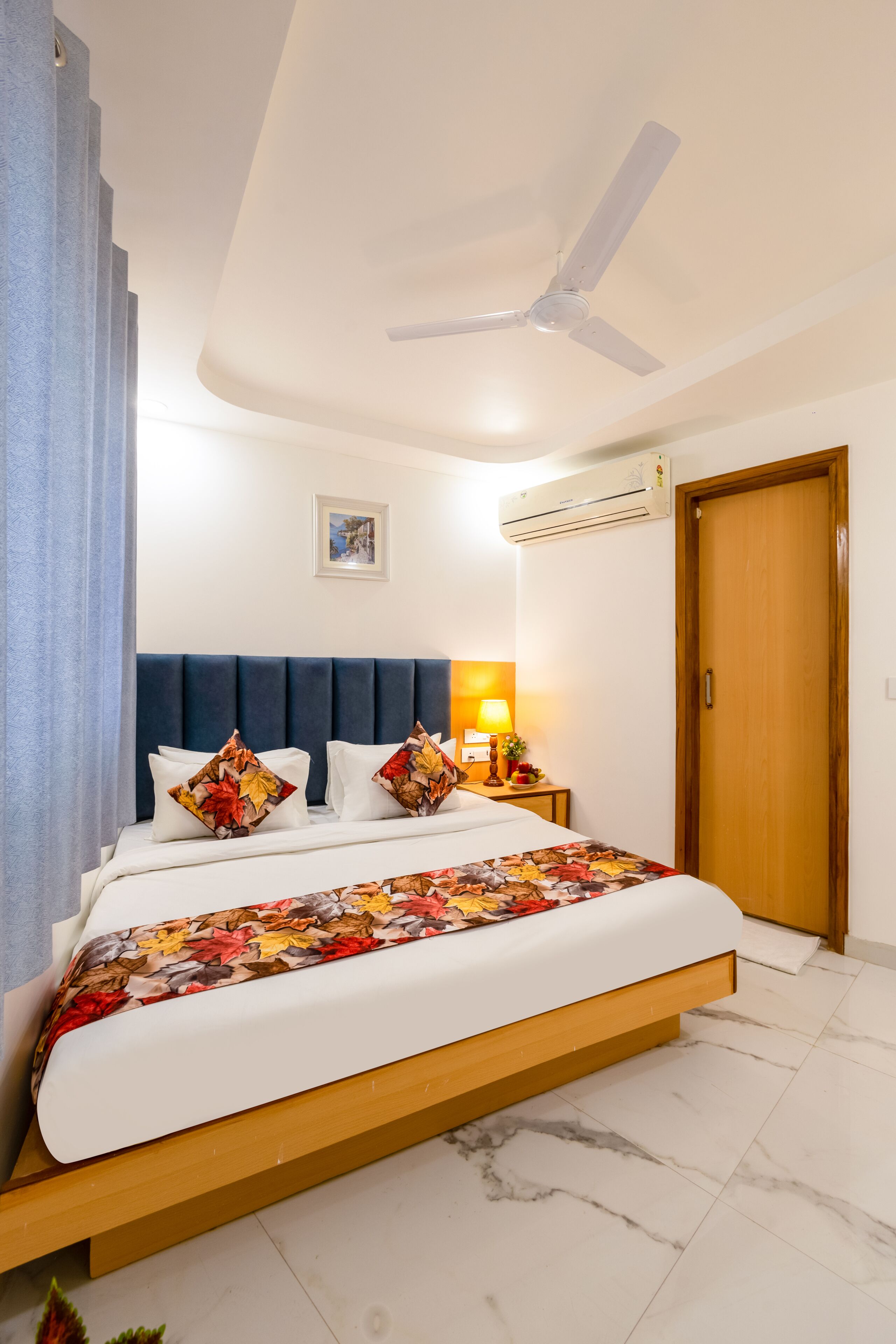 A bedroom with a bed, a ceiling fan, an air conditioning unit, a wooden door, and a nightstand with a lamp.