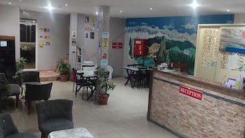 Un área de recepción con mostrador, sillas, mesas, plantas en macetas y un mural en la pared.