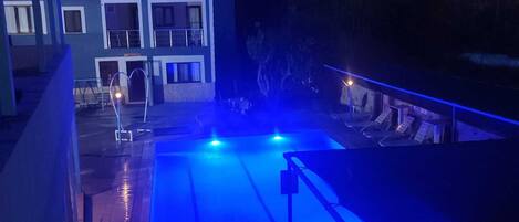 A swimming pool with blue lighting, a building with balconies, and a view of trees.