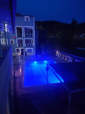 A swimming pool with blue lighting, a building with balconies, and a view of trees. - Anatolia Hotel (Denizli)