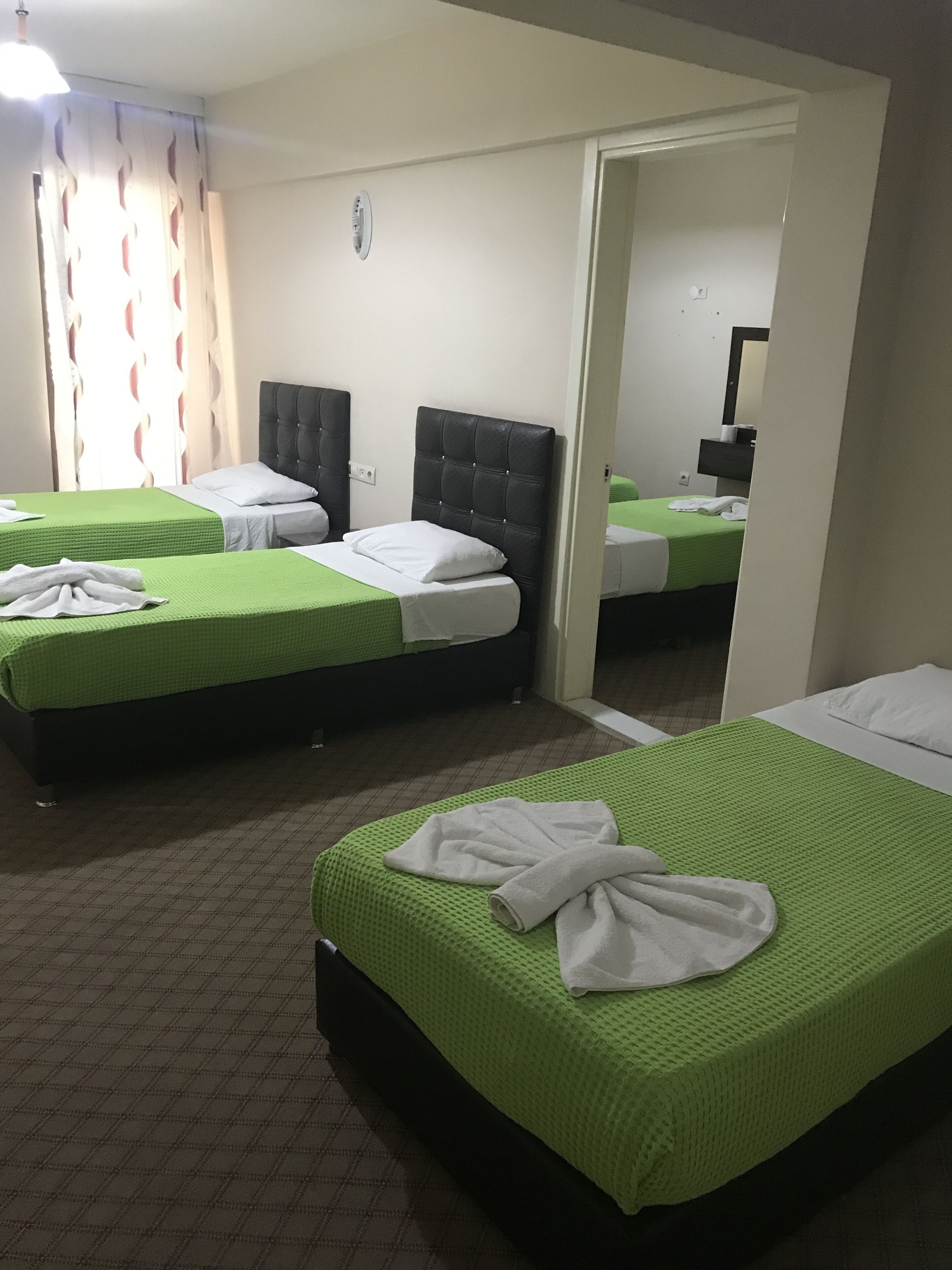 A hotel room with two beds, each with a green bedspread and white pillows. There is a mirror on the wall and a window with curtains.