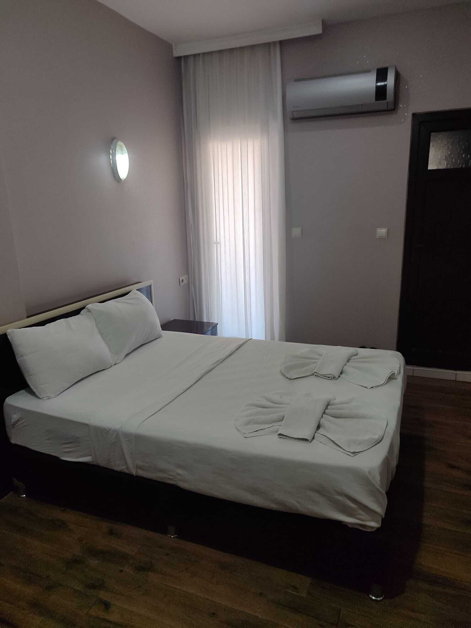 A hotel room with a bed, a bedside table, a window with curtains, and an air conditioning unit on the wall.