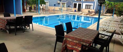 A poolside area with lounge chairs and tables, a clear blue pool, and a building with balconies in the background.