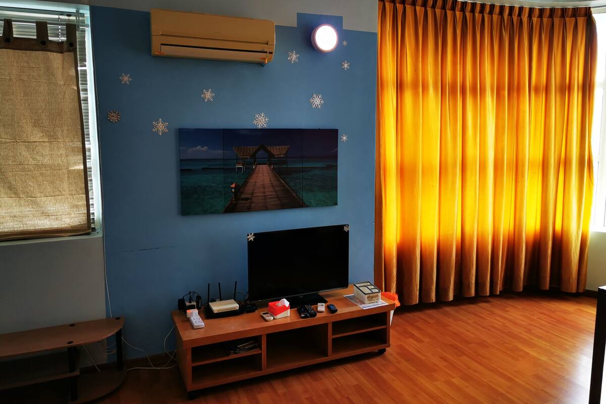 Une pièce dotée d’un plancher en bois, d’un climatiseur fixé au mur, d’un téléviseur à écran plat, d’un banc en bois, d’une table avec des appareils électroniques et d’une grande fenêtre avec des rideaux.