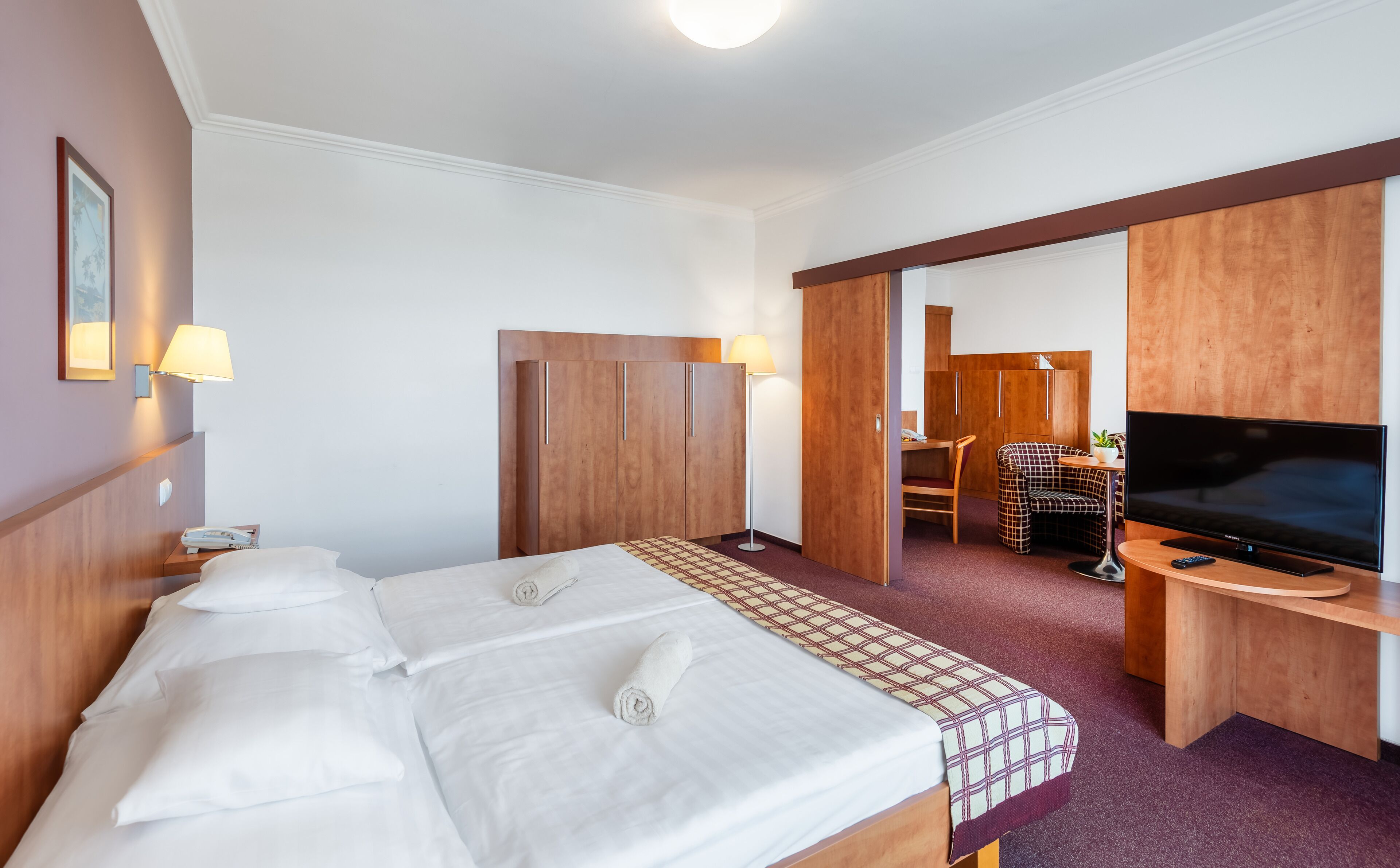 A hotel room with a large bed, a desk with a TV, a chair, and a wardrobe.