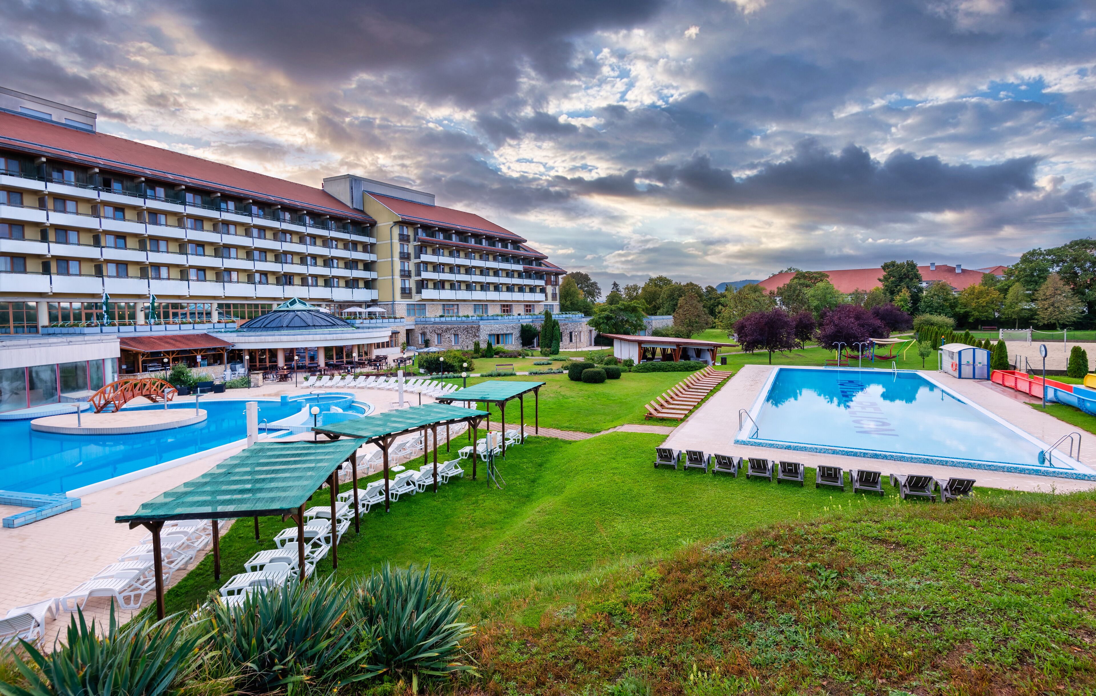 A hotel with multiple swimming pools, a covered seating area, and a well-maintained lawn.