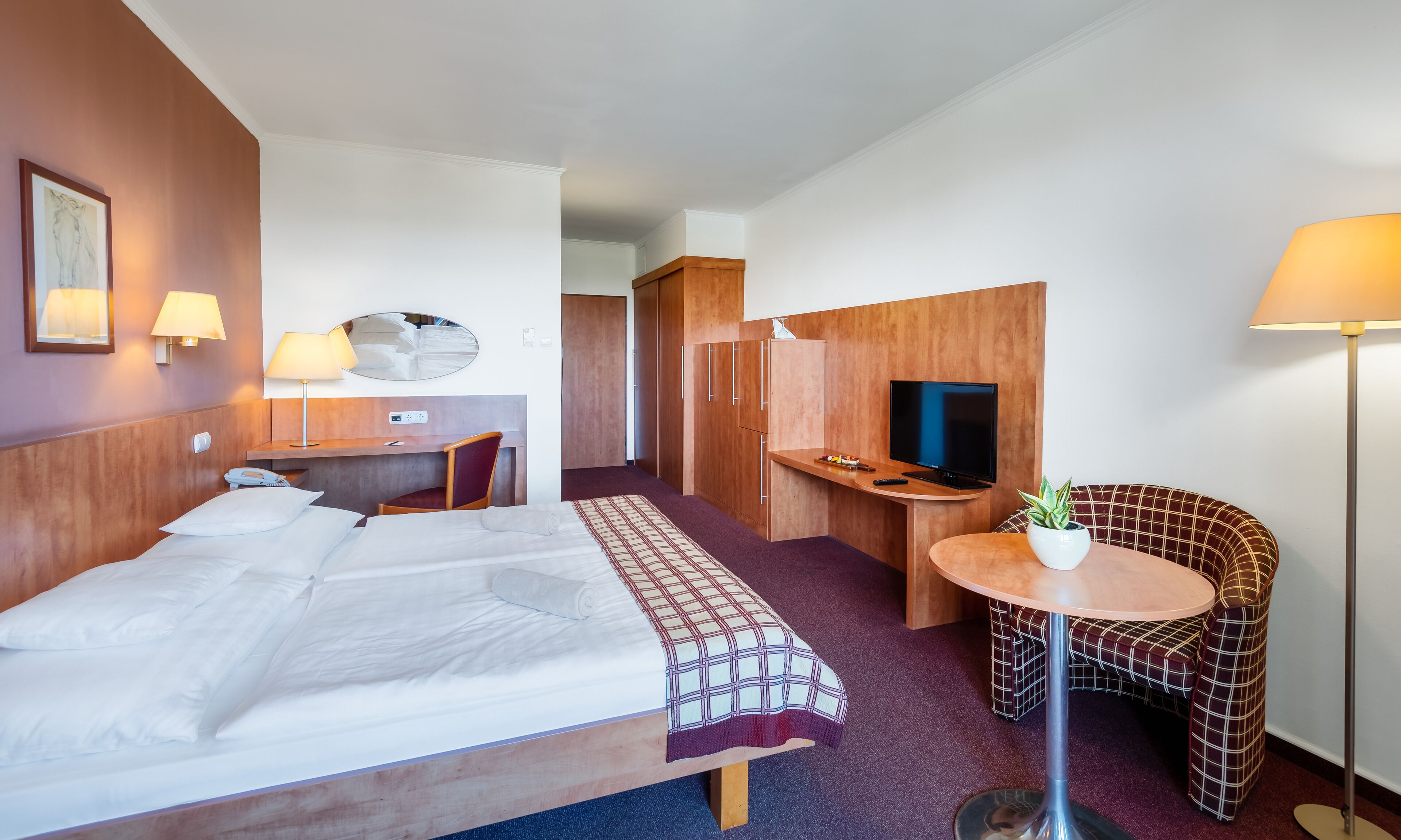A hotel room with a bed, desk, chair, TV, and a small table with a plant.