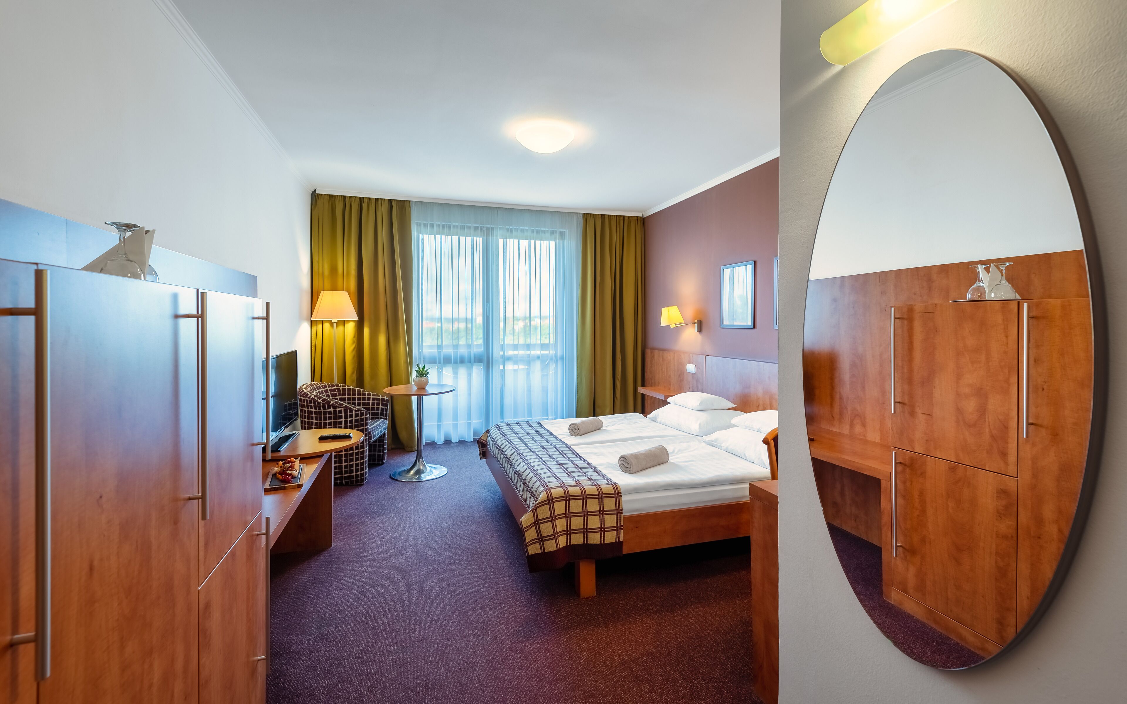 Habitación de hotel con cama, sofá, escritorio, espejo y vistas al exterior.