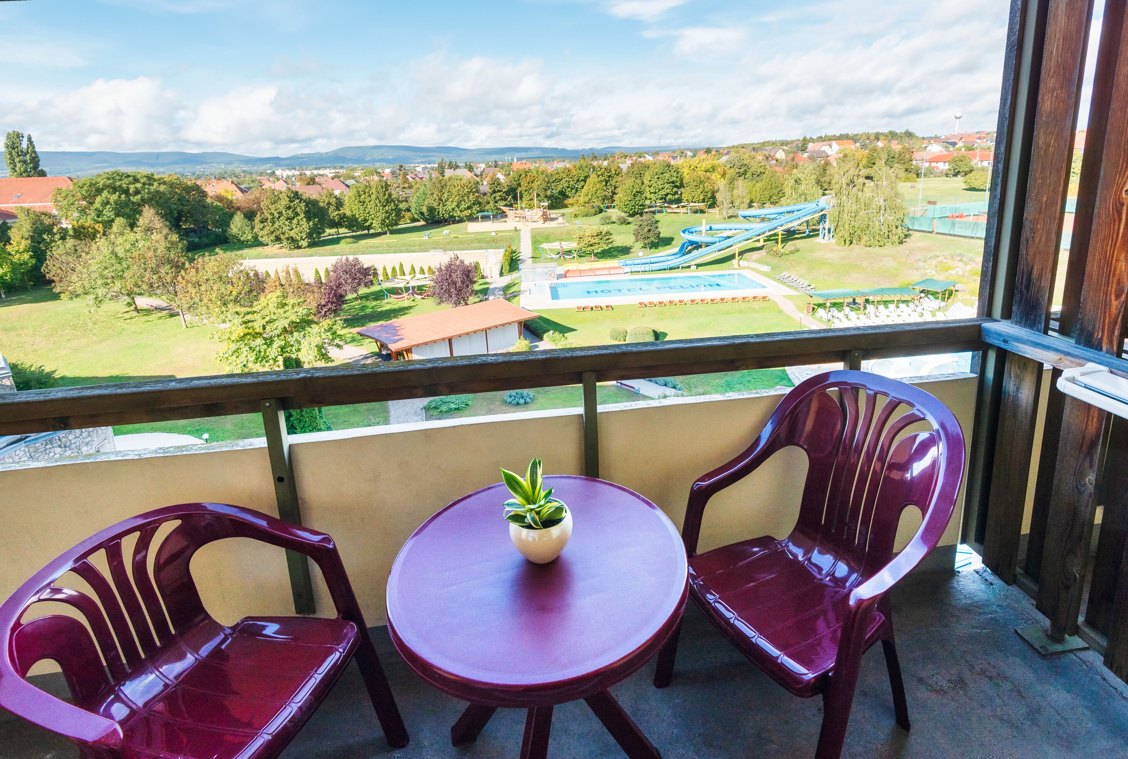 A balcony with two plastic chairs and a small table with a potted plant. A view of a hotel complex with a pool and a water slide is visible in the background.