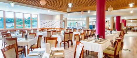 A spacious dining area with multiple tables and chairs, large windows, and a wooden ceiling with a grid pattern.