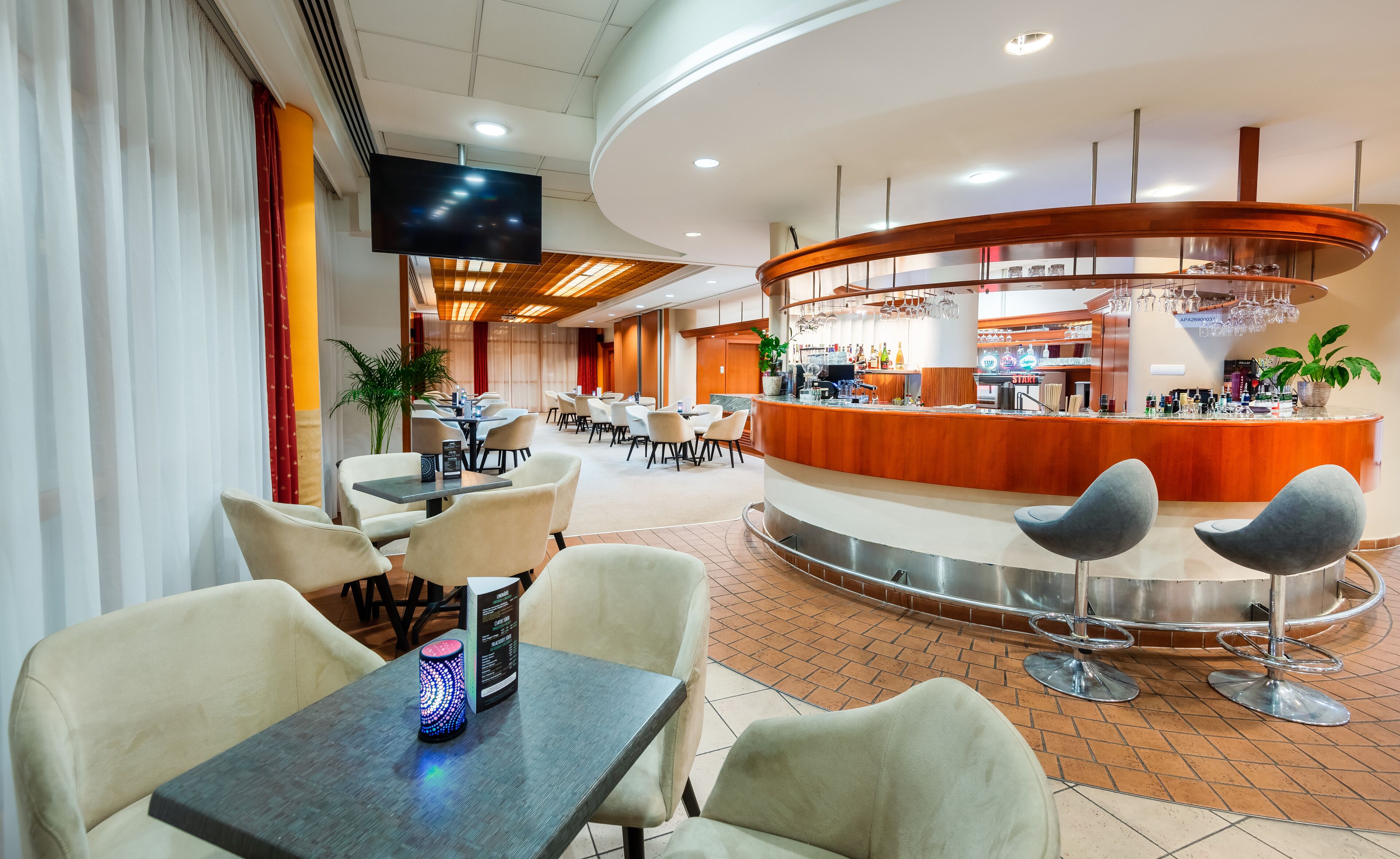 A modern hotel lobby with a central bar area, multiple seating arrangements, and a flat-screen TV mounted on the wall.