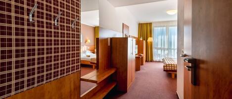 A hotel room with a wooden wardrobe, a bed with a plaid bedspread, a small desk with a lamp, and a window with sheer curtains.
