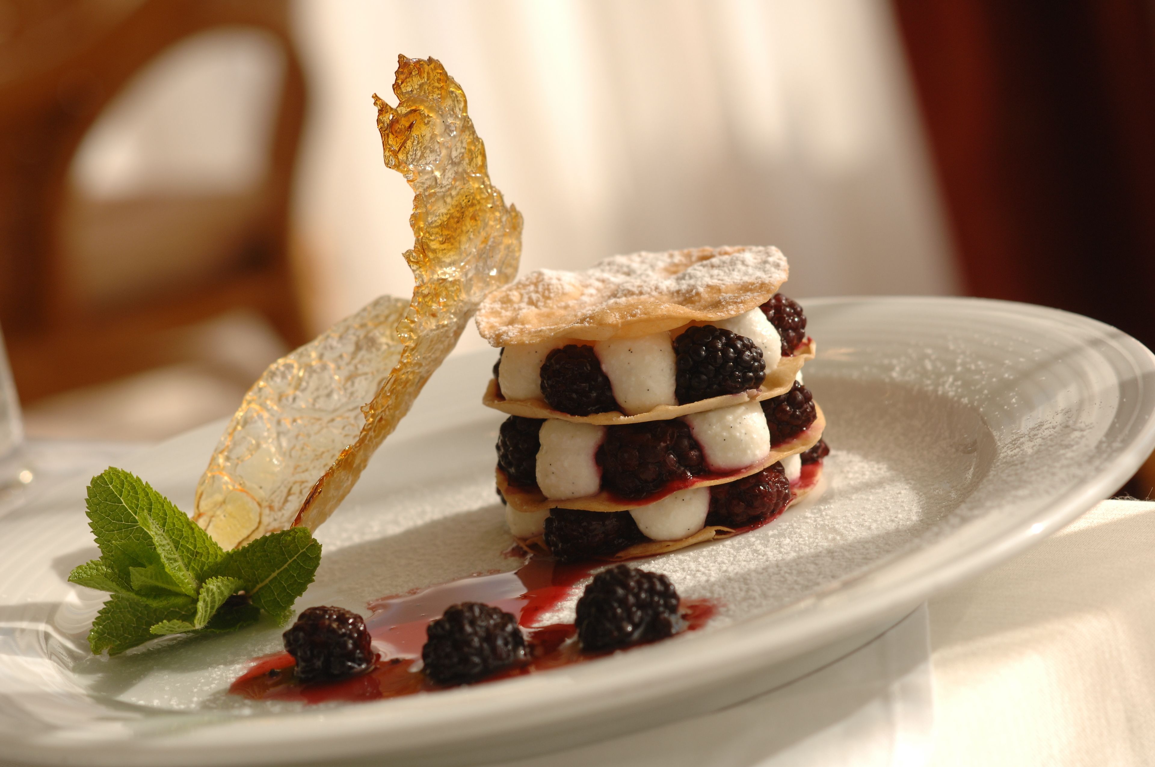 Postre con capas de frutos rojos y crema, espolvoreado con azúcar glas, acompañado de un trozo de hojaldre crujiente y una hoja de menta sobre un plato blanco.