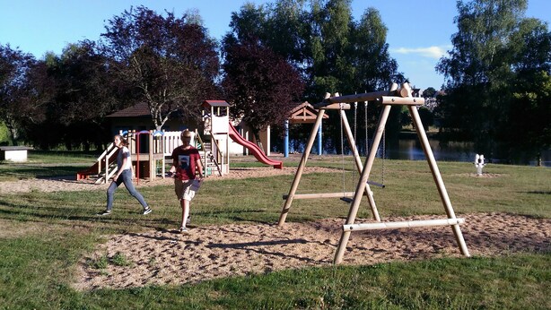 A playground with swings and a slide, surrounded by trees and a lake.