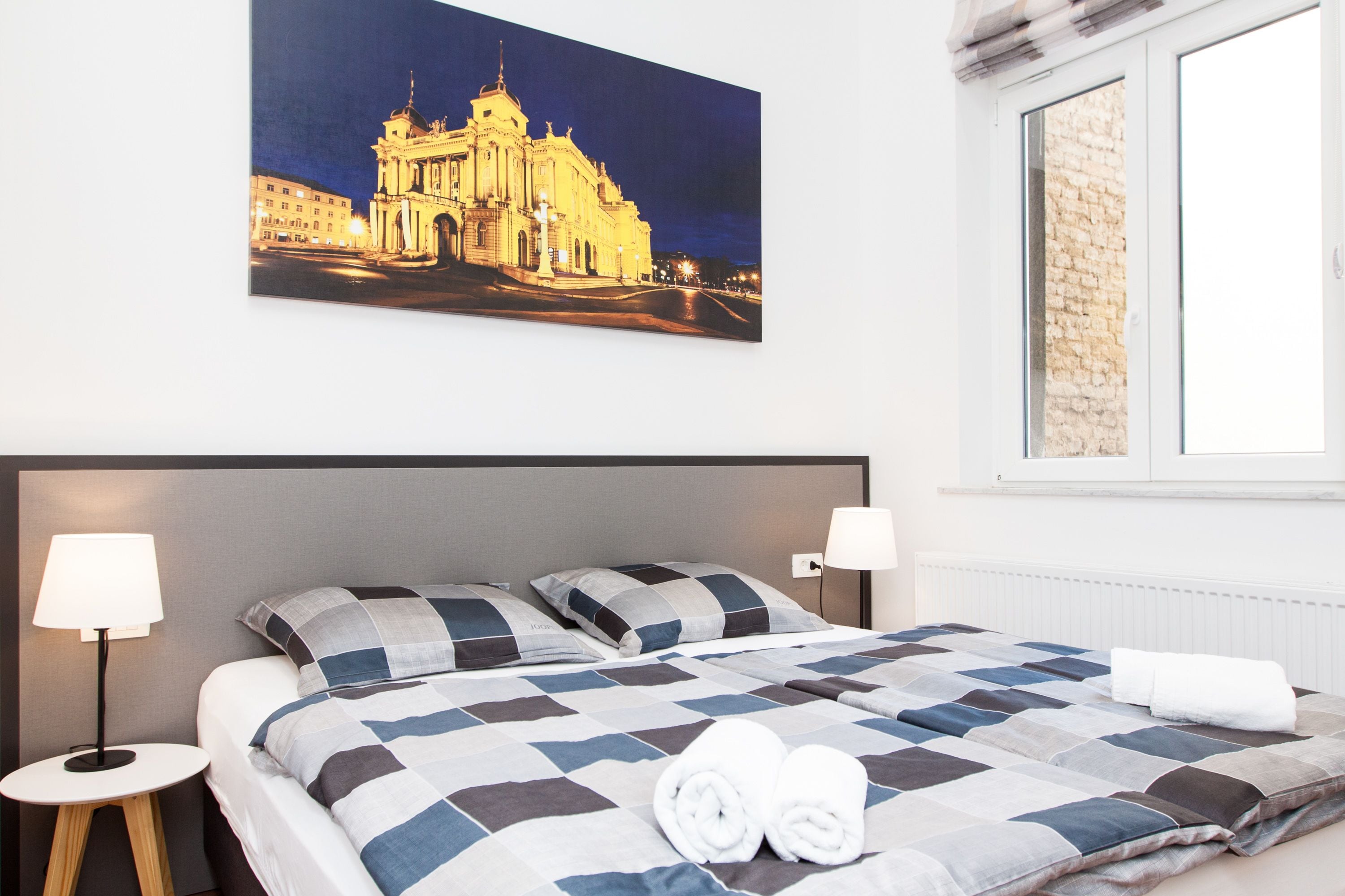 A hotel room with a large bed, two bedside lamps, a window with a brick wall, and a framed picture of a building on the wall.