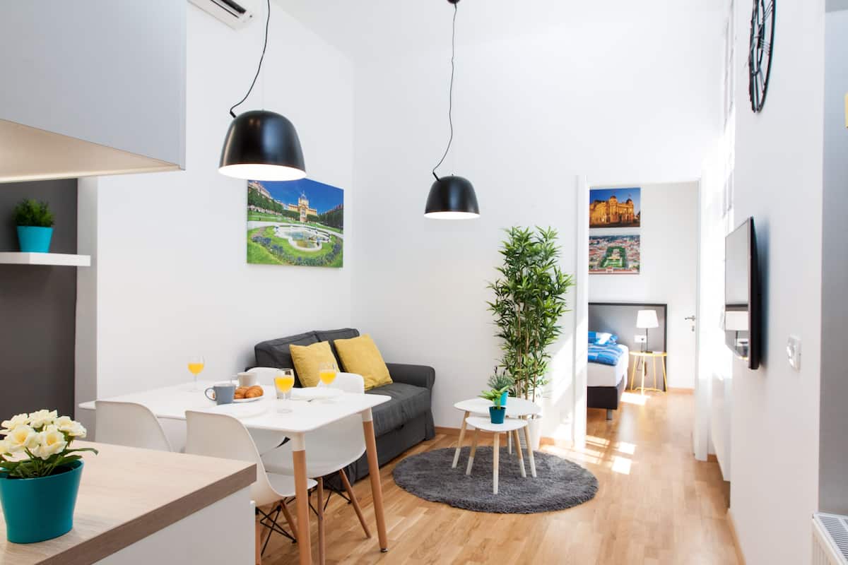 Une pièce moderne et bien éclairée, comprenant un coin repas, un canapé, une petite table avec une plante et un lit à l’arrière-plan.