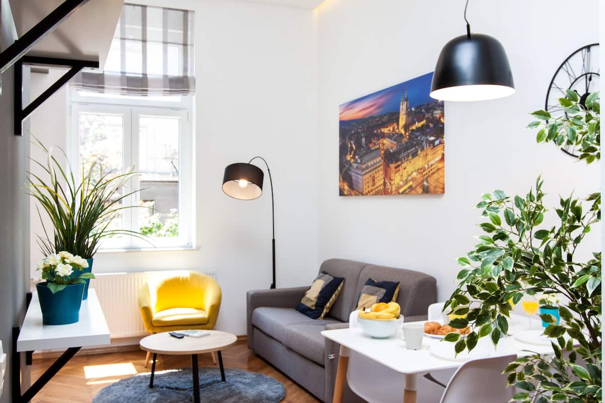Un salon moderne avec un canapé, un fauteuil jaune, une table à manger avec des aliments pour le petit-déjeuner, et un tableau représentant un paysage urbain accroché au mur.