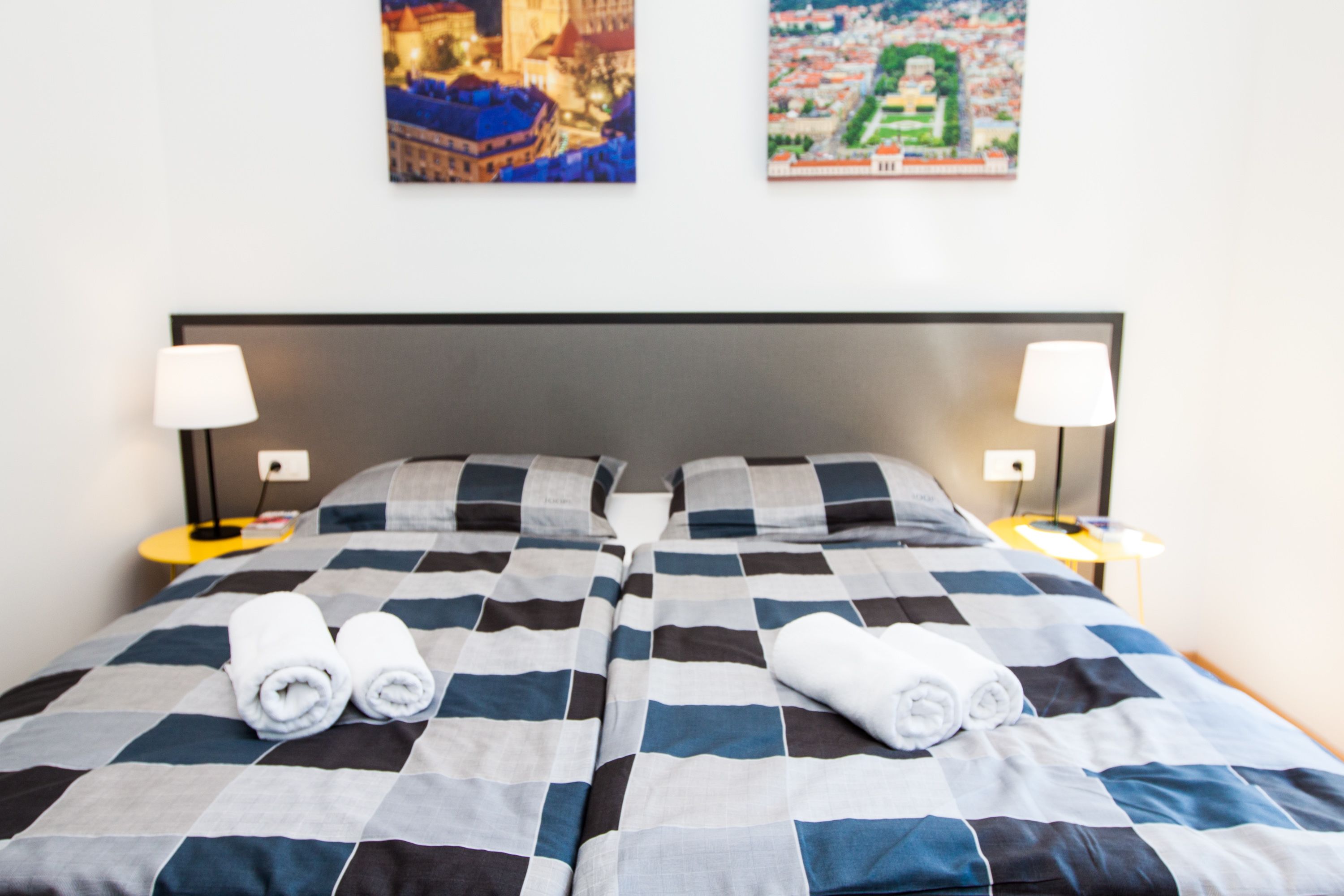 A double bed with a checkered bedspread, two pillows, two bedside lamps, and two rolled towels on the bed.