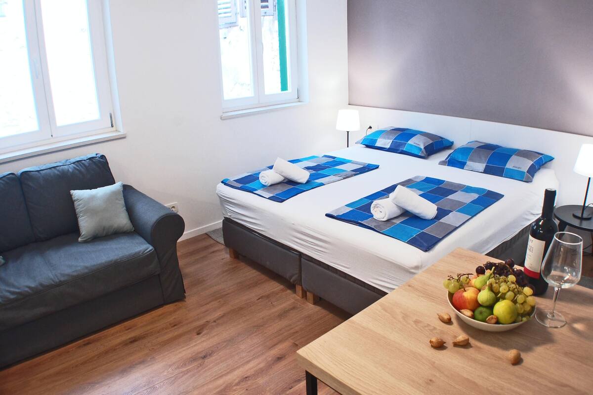 Une chambre d’hôtel avec un lit, un canapé, une table en bois sur laquelle se trouvent des fruits et une bouteille de vin, et une fenêtre.
