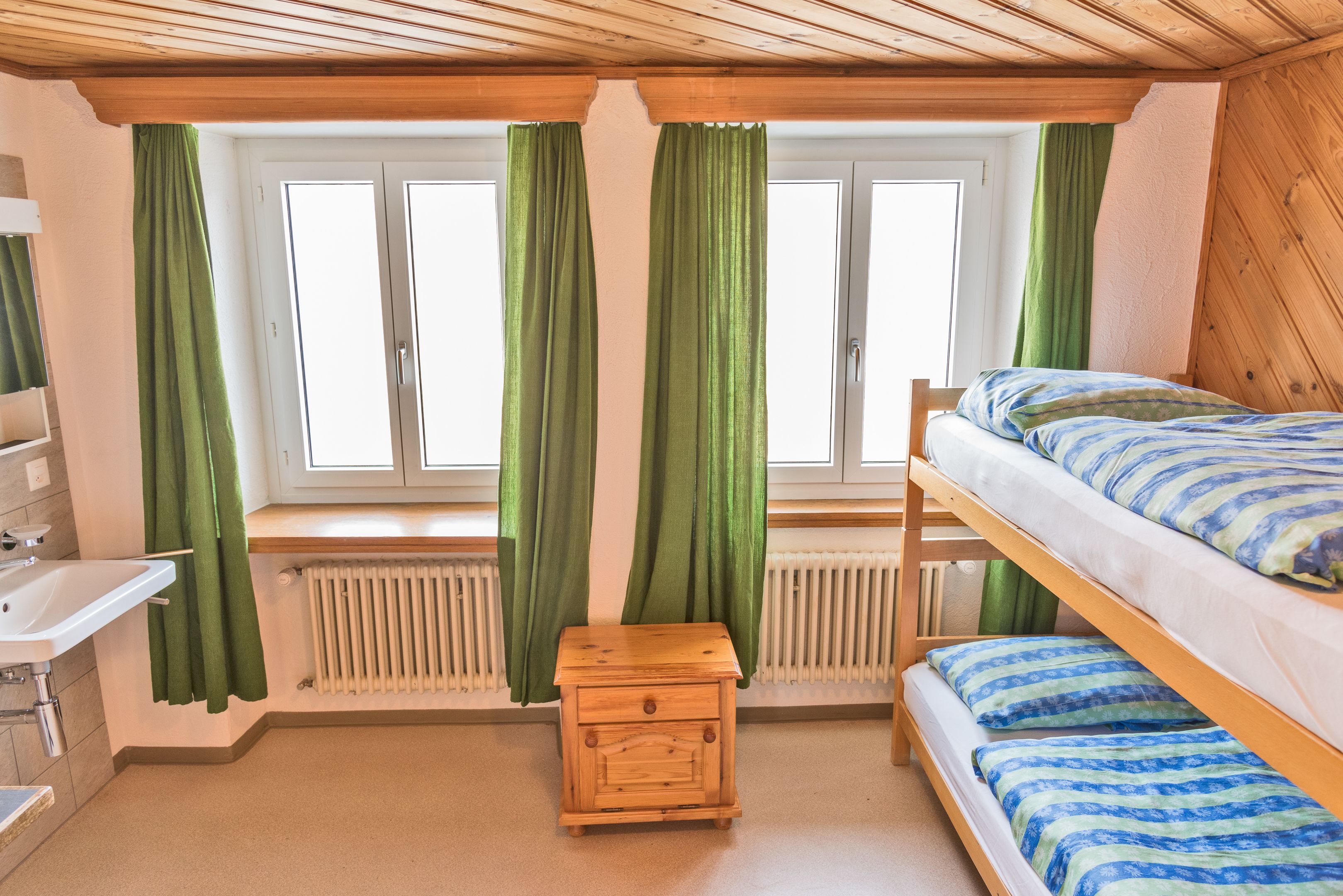 A small, cozy room with a bunk bed, a wooden nightstand, a white sink, and a window with green curtains.