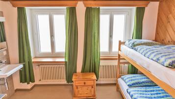 A small, cozy room with a bunk bed, a wooden nightstand, a white sink, and a window with green curtains.