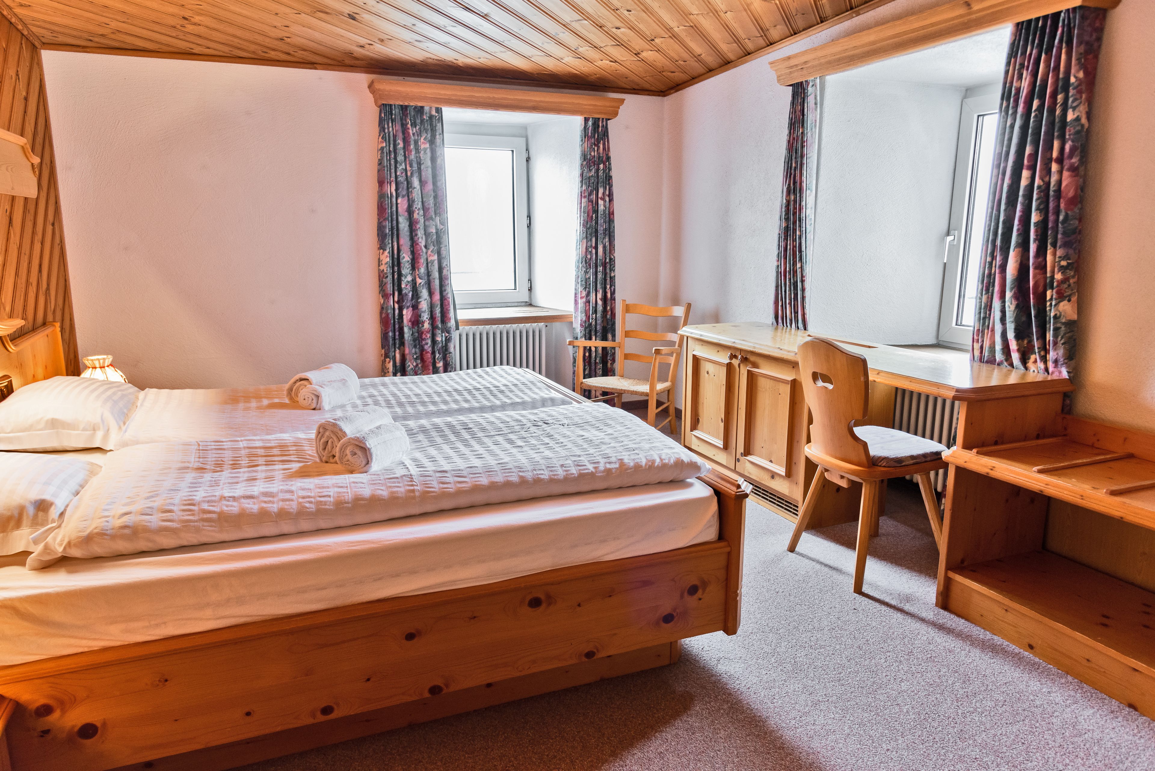 A wooden bed with a mattress and pillows, a wooden desk with a chair, and a window with curtains.