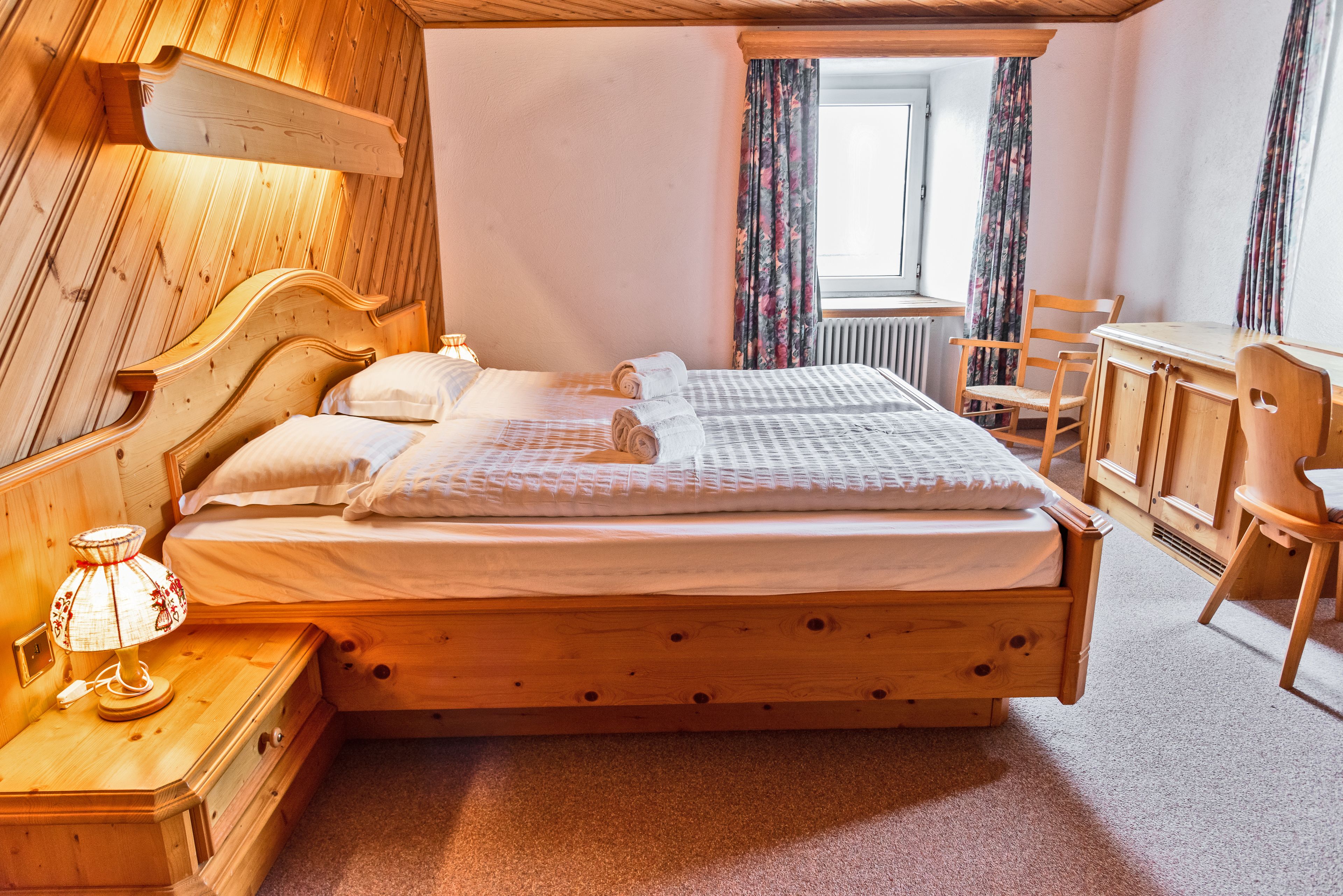 Un lit en bois avec une tête de lit, une table de chevet avec une lampe, un bureau avec des chaises et une fenêtre avec des rideaux.